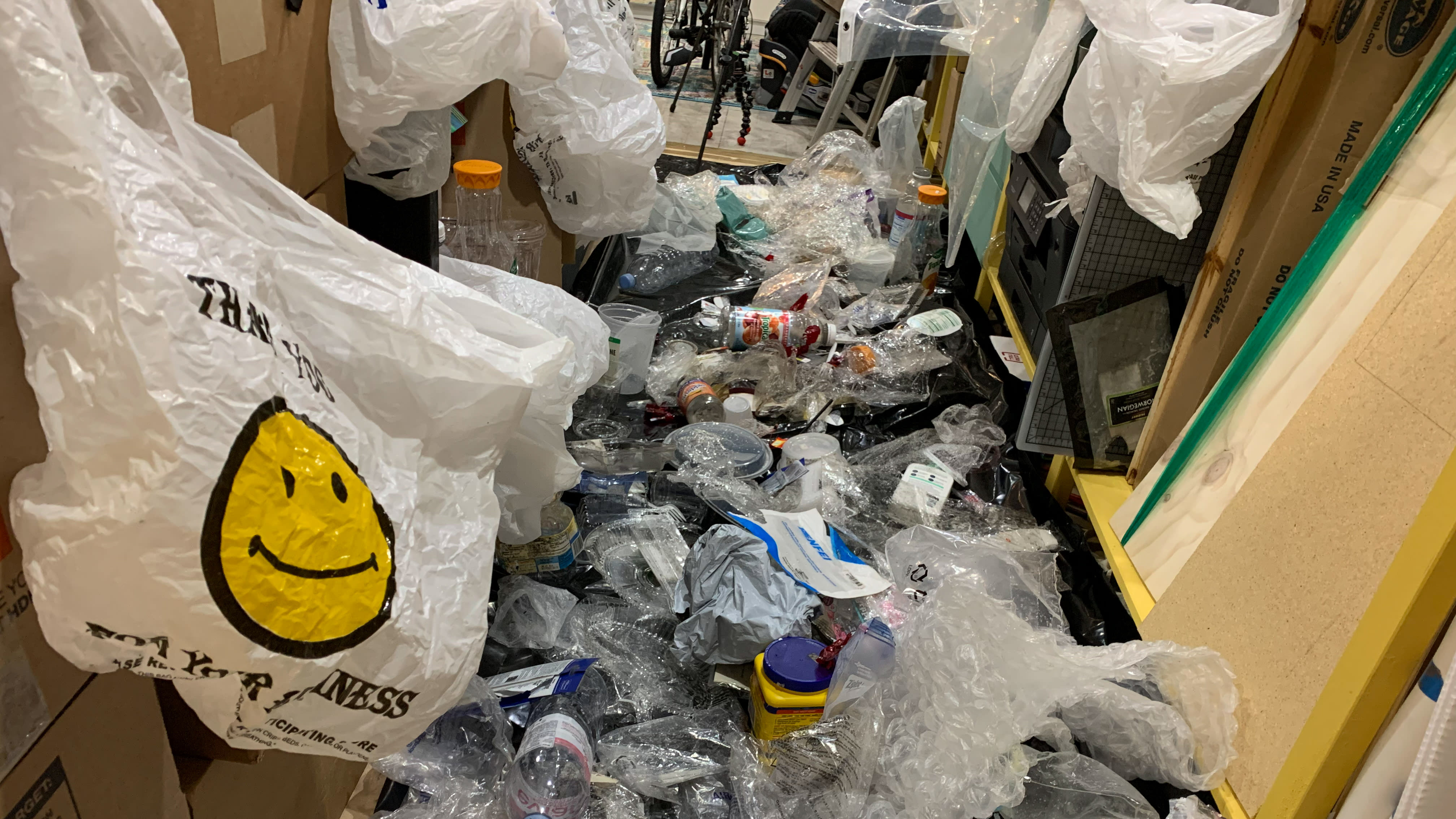 Photograph of the artist's studio. On the left of the frame is a close-up of a white plastic 'Thank You' shopping bag with a smiley face on it. The middle and right of the frame show hundreds of sundry single-use plastic items strewn about the floor.