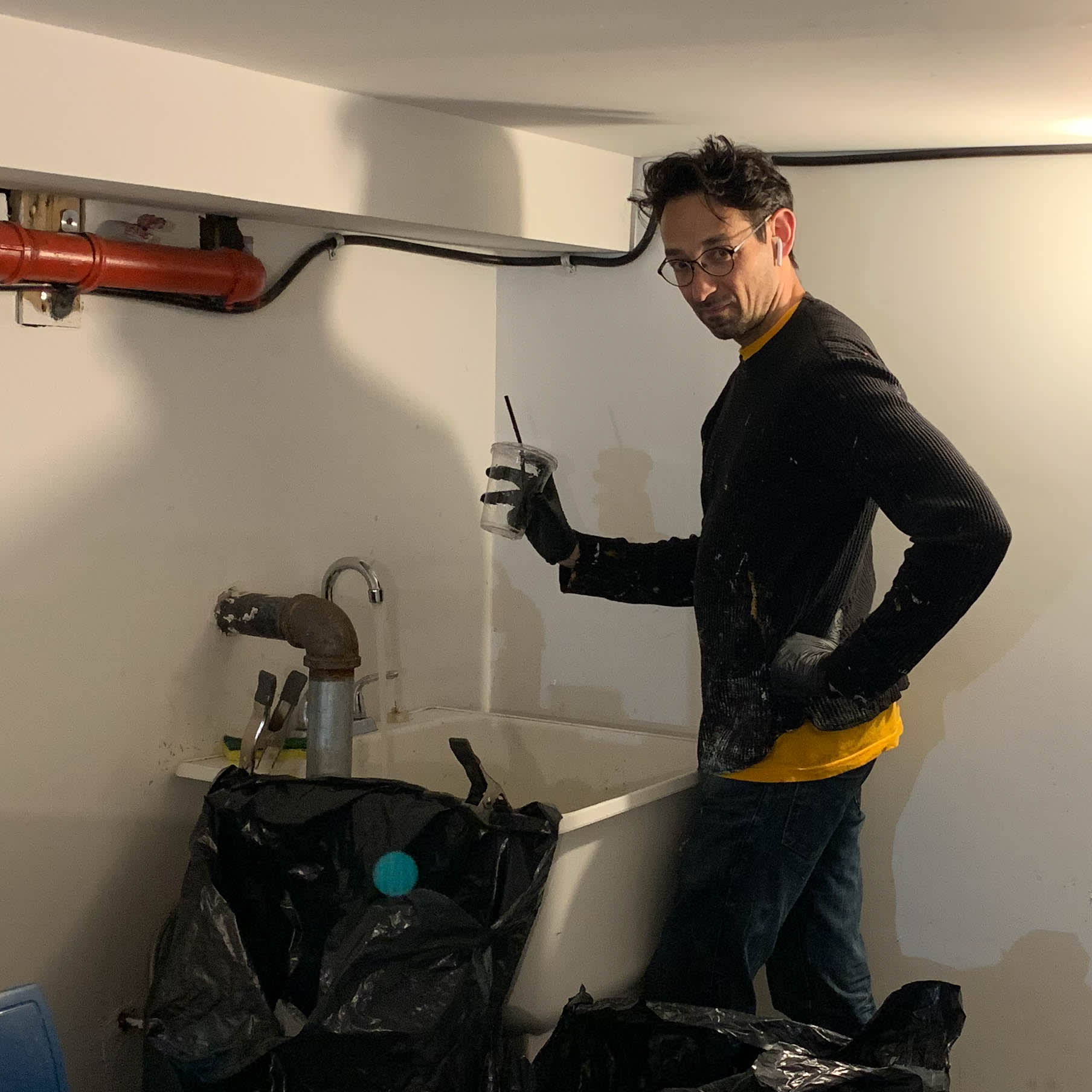 Photograph of a man (the artist), looking at the camera and dressed in tattered, 'art studio' wear, standing by a laundry sink (in a basement) holding a plastic cup as though he is about to wash it.