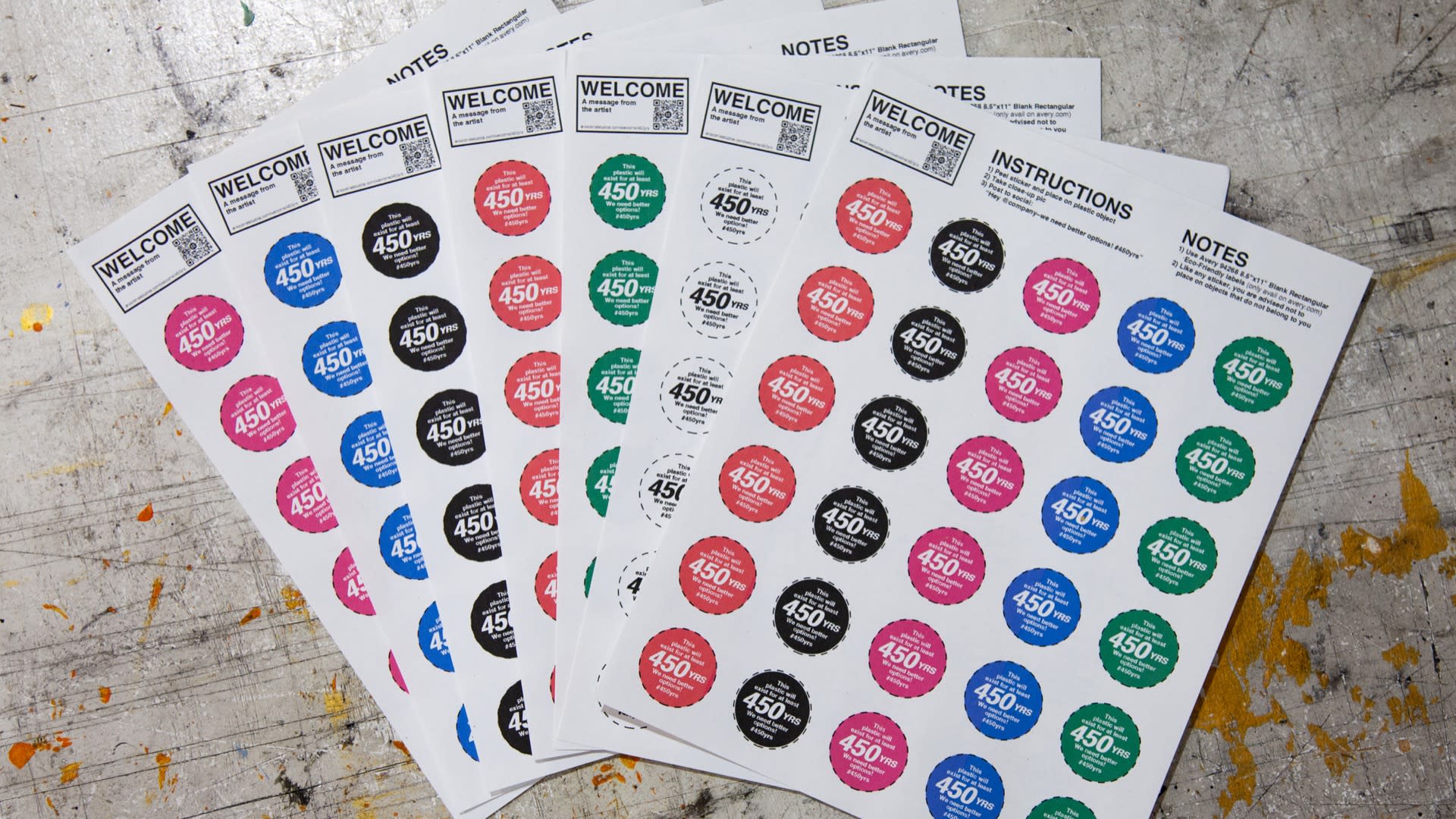 Photograph of seven 8.5 inch by 11 inch pieces of paper, laid out in a fan-like fashion, on a workbench. Each sheet shows contains 30 DIY, approximately 1.5 inch diameter stickers. Individual stickers are circular in nature, come in either red, green, blue, magenta, black, and white and the all say the same thing: 'This plastic will exist for at least 450YRS. We need better options! #450yrs '