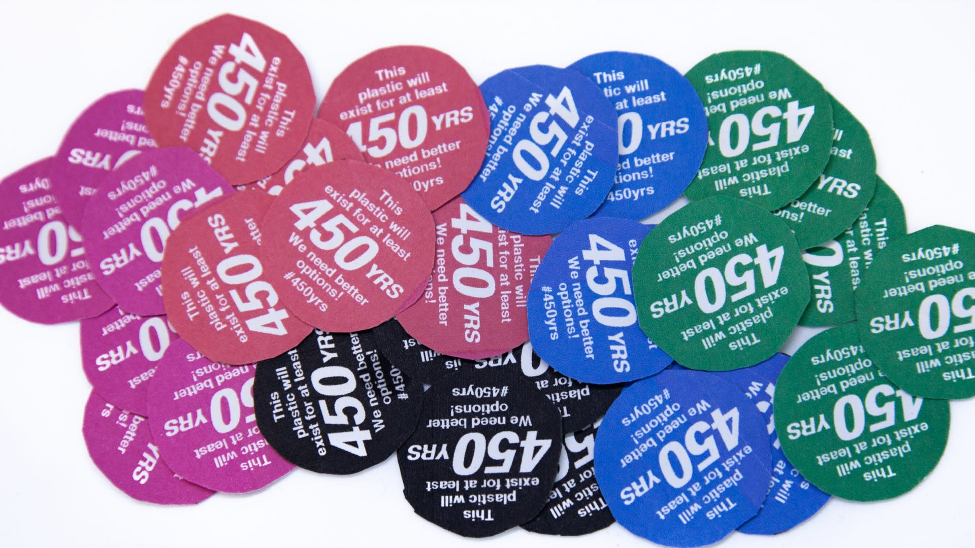 Thumbnail of Close-up photograph of approximately 30, hand-cut out, circular stickers–approximately 1.5 inches in diameter, all bunched up together on a white background. Stickers are either red, green, blue, magenta, black, or white and all say 'This plastic will exist for at least 450YRS. We need better options! #450yrs '
