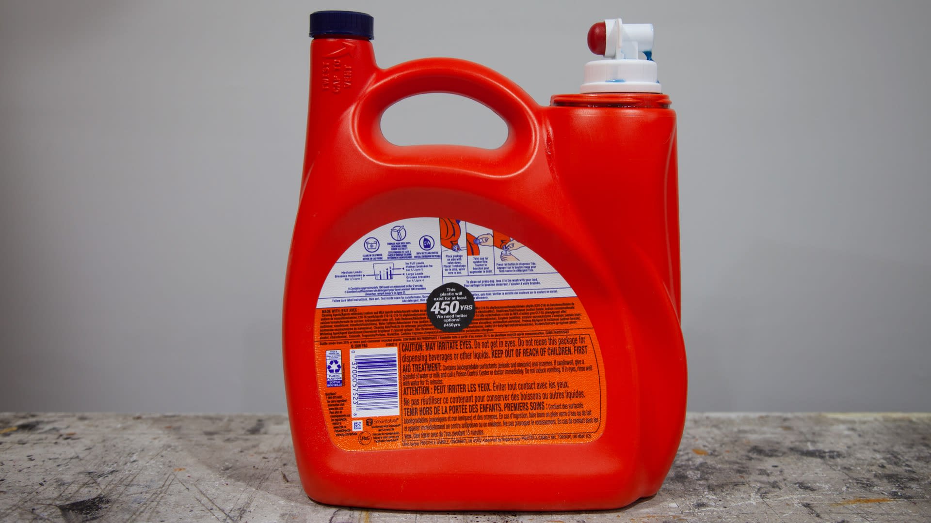 Thumbnail of Photograph of a large, red, plastic laundry detergent bottle sitting on a workbench. In the middle of the bottle is a 1.5 inch, circular sticker that says, 'This plastic will exist for at least 450YRS. We need better options! #450yrs '