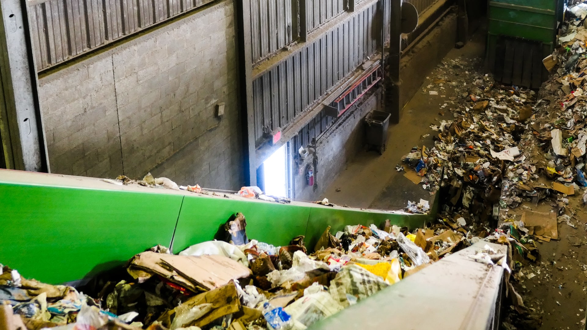 Photo of the interior of an industrial recycling facility. The left side of the photo is dominated by a large conveyor belt that is bringing trash up from ground level, about 20 feet below.