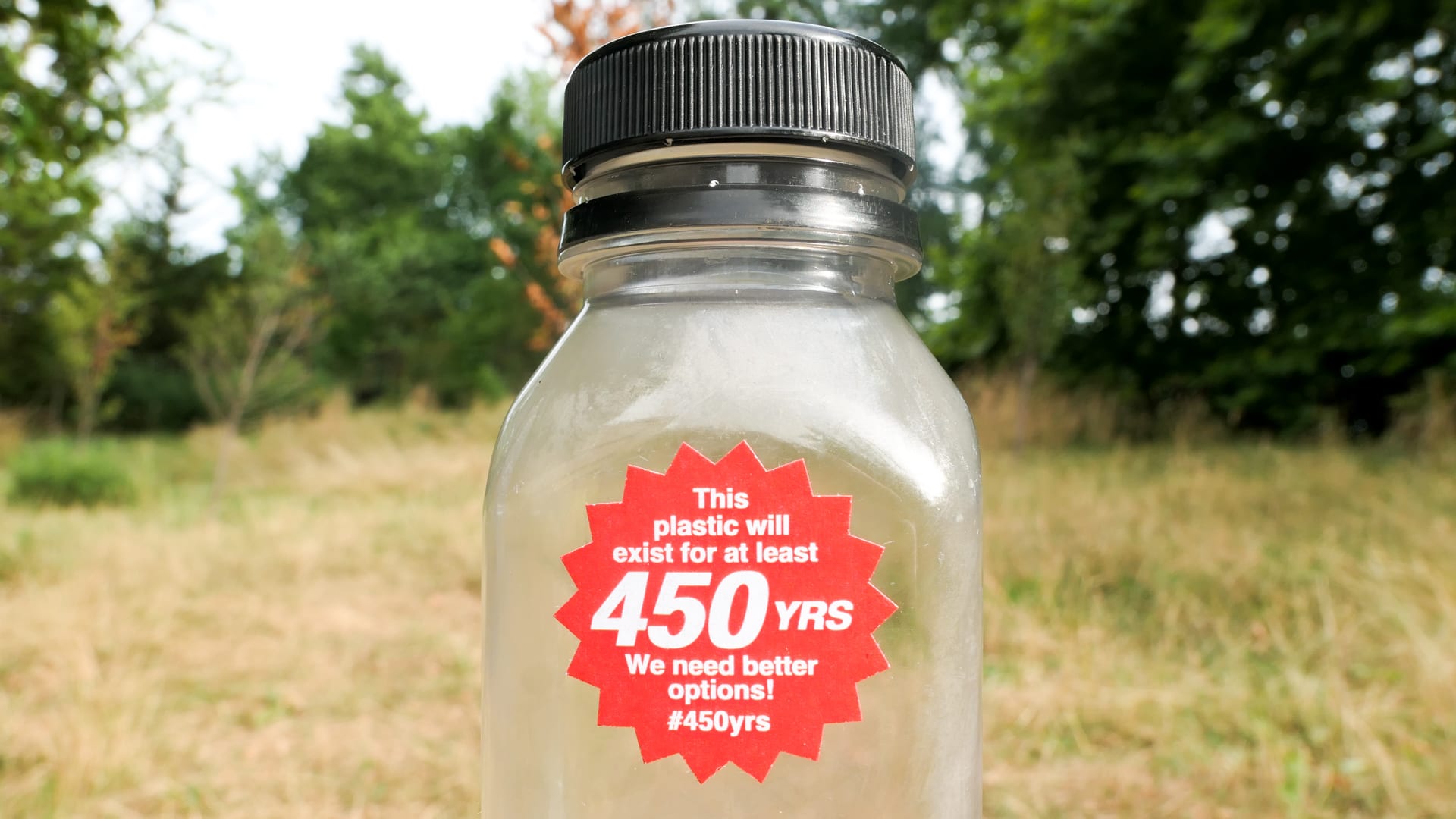 Photograph of a single-use plastic bottle with a red, starburst-shaped 1.25 inch sticker on it that says: 'This plastic will exist for at least 450 years.
        We need better options #450yrs.' In the background, behind the bottle, is a green field.