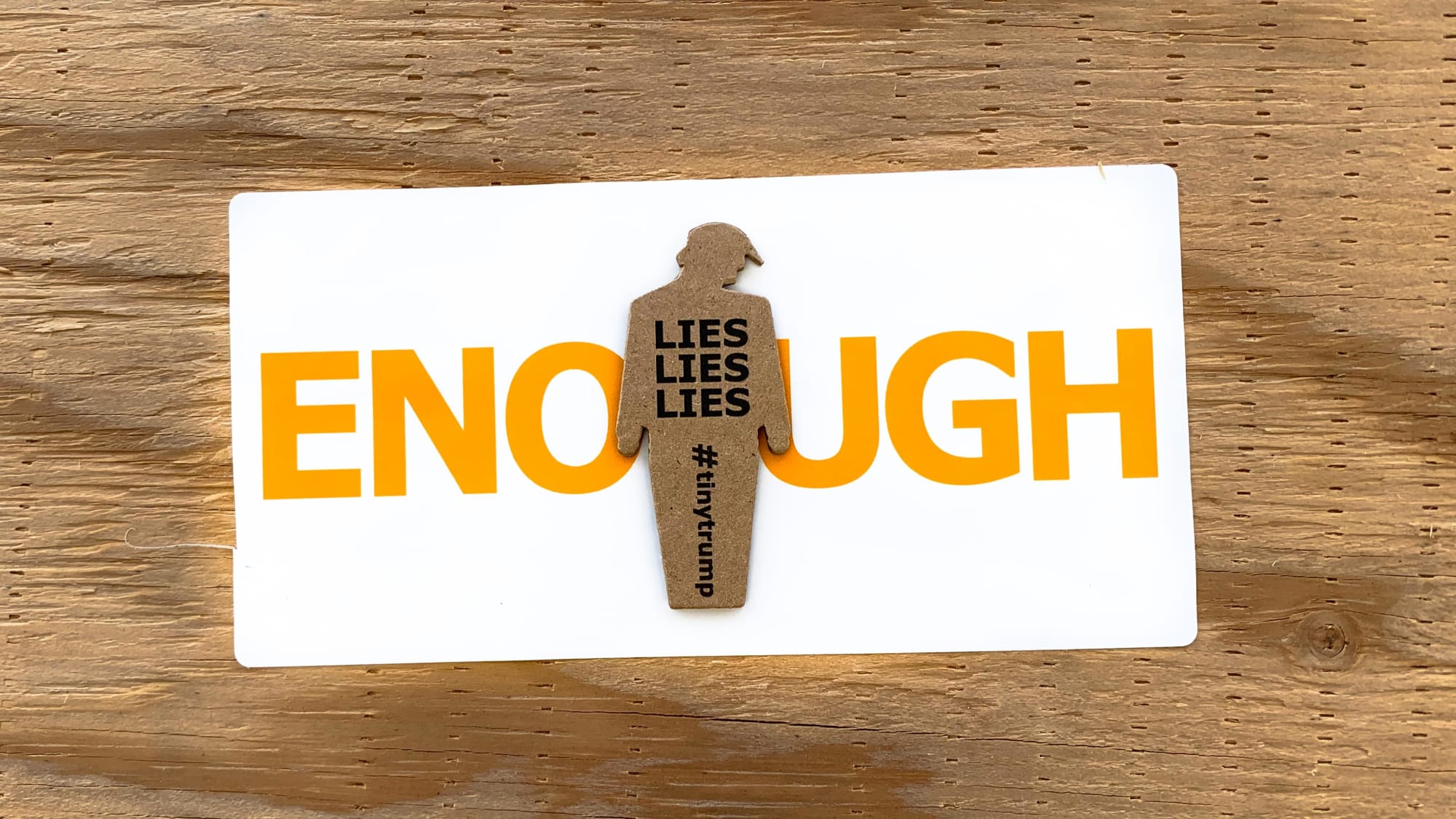 Photograph of a tiny trump with the slogan Lies Lies Lies stuck in the middle of a white bumper sticker with large orange type that says ENOUGH in all caps