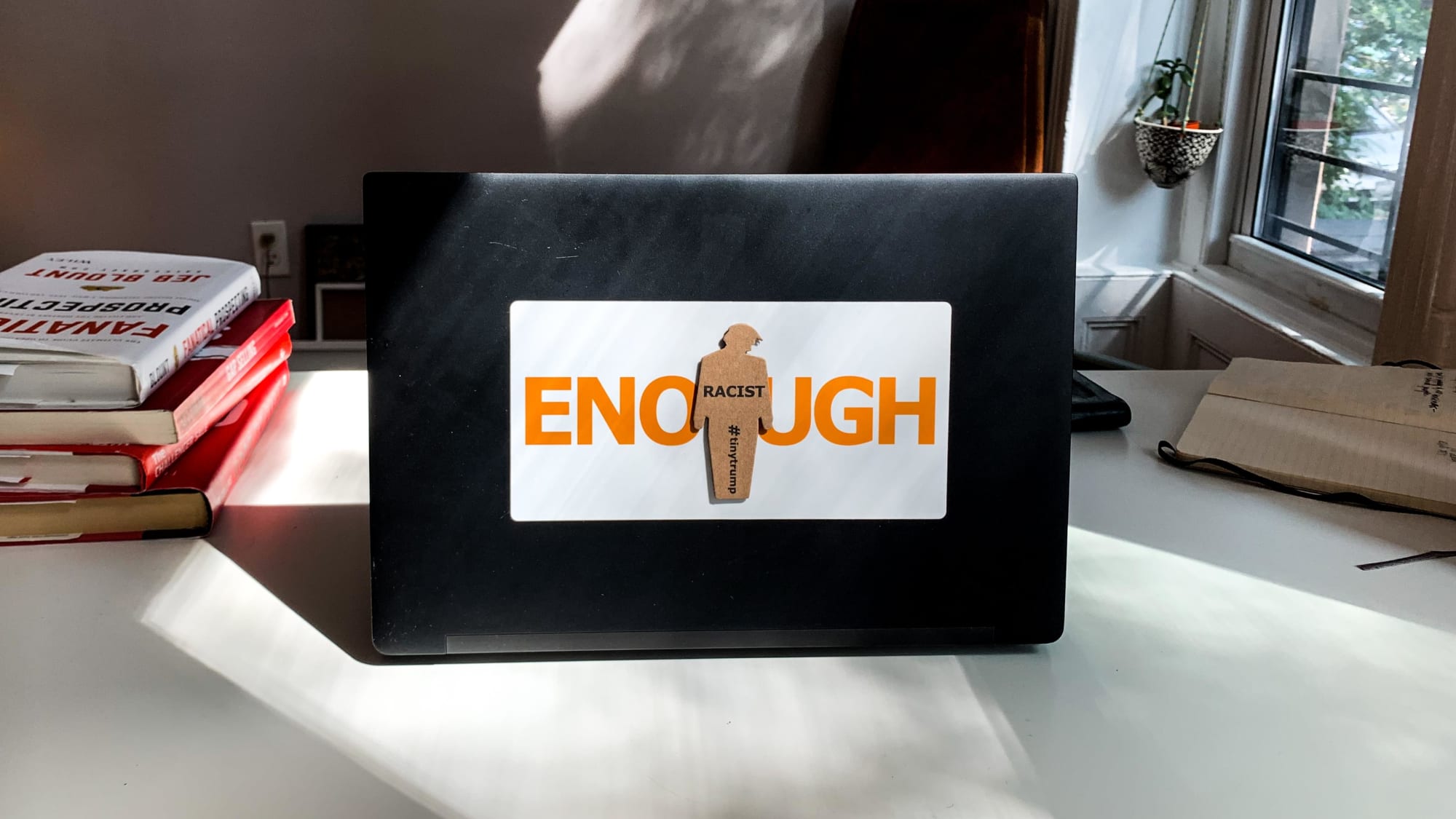 Photograph of a tiny trump ENOUGH sticker on a laptop. The tiny trump stuck in the middle bears the slogan Racist, so the combination reads ENOUGH Racist