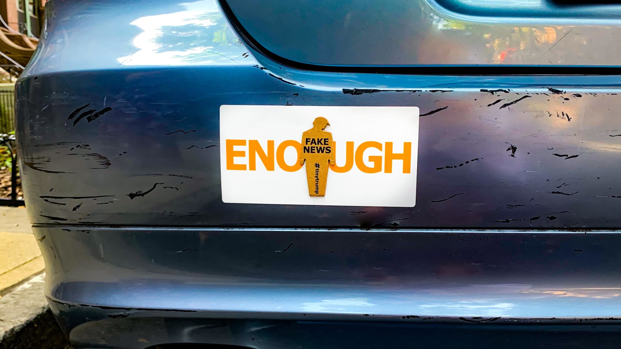 Photograph of a tiny trump ENOUGH sticker on a car bumper. The tiny trump stuck in the middle bears the slogan Fake News, so the combination reads ENOUGH Fake News.