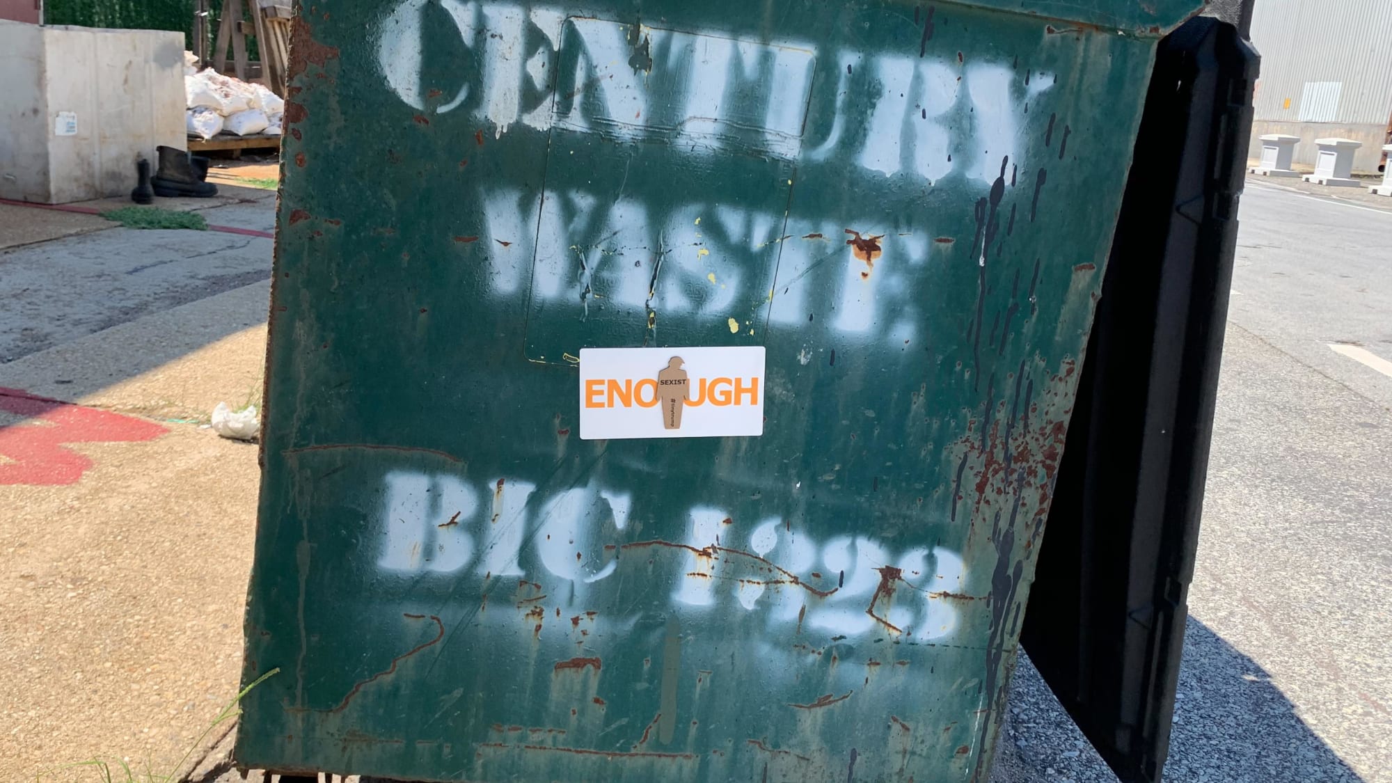 Photograph of a tiny trump ENOUGH sticker on a dumpster in the street. The tiny trump stuck in the middle bears the slogan Sexist, so the combination reads ENOUGH Sexist