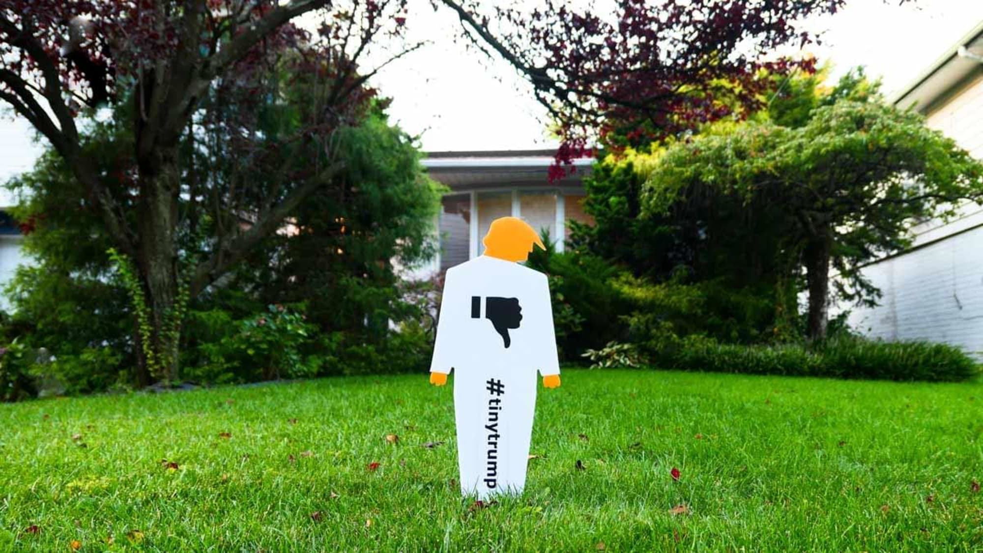 Two foot tall lawn sign in the shape of a tiny trump—a cardboard cutout caricature of trump—with a thumbs down sign on the chest. The lawn sign is made out of plastic, is white with orange head and hands. It is mounted on a lawn in front of a suburban house which is in the background.