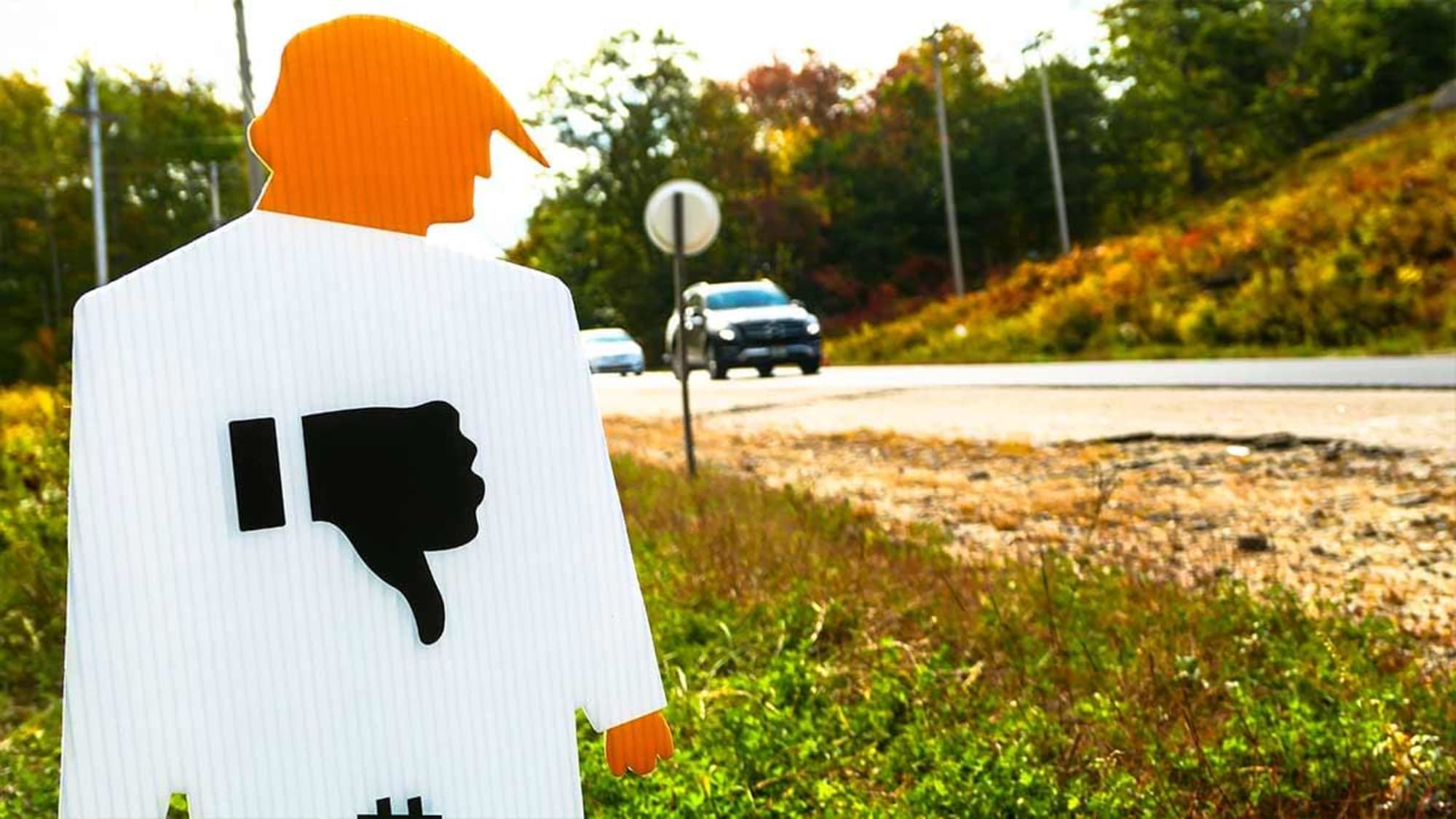 Close up photograph of a tiny trump lawn sign shown on the side of a road. A blurred SUV on a road is in motion in the background.