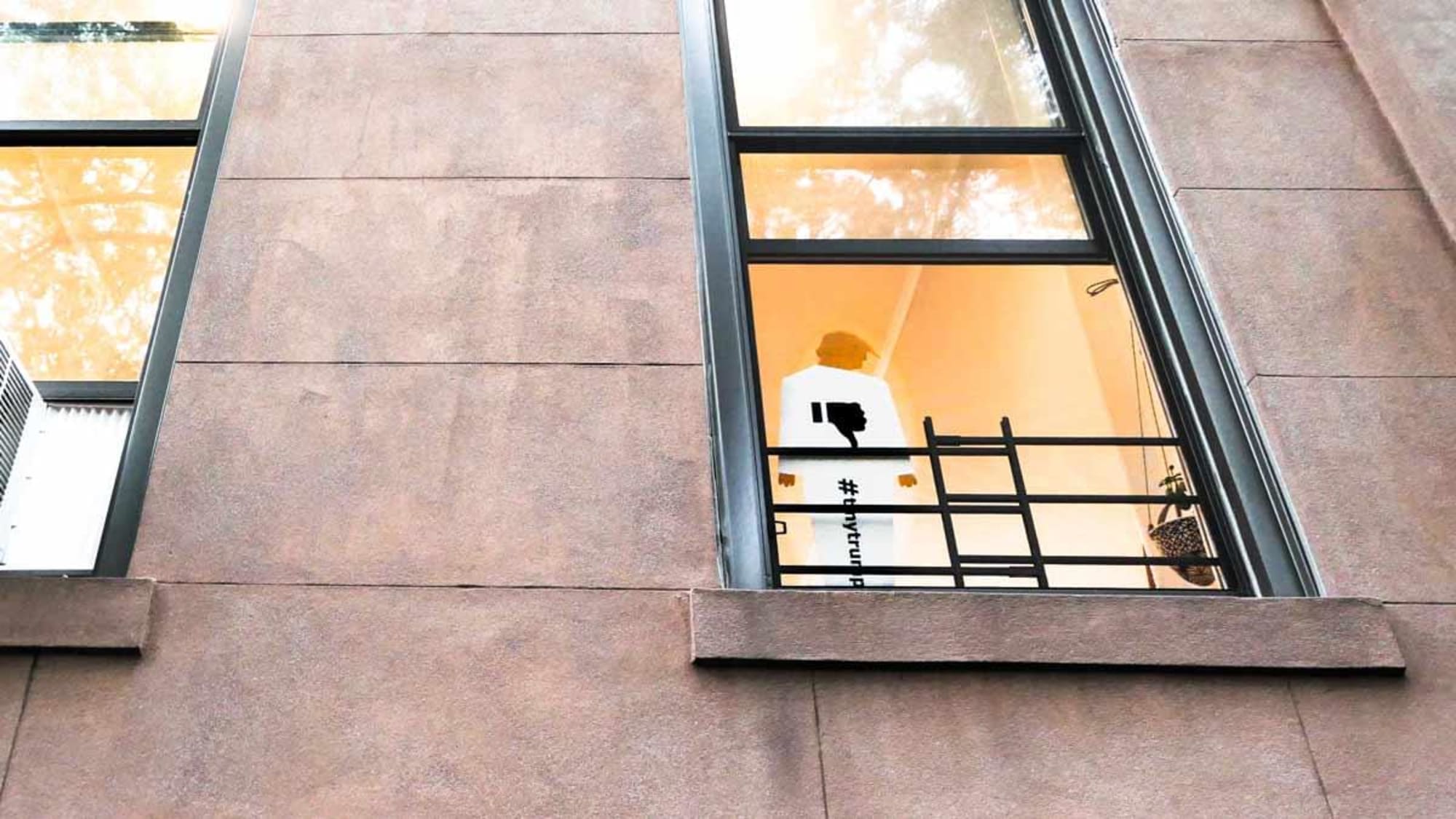 Photograph of a tiny trump lawn shown in an apartment window. The perspective of the photograph is sharply upward, with the tiny trump occuping the lower half of a brownstone apartment window.