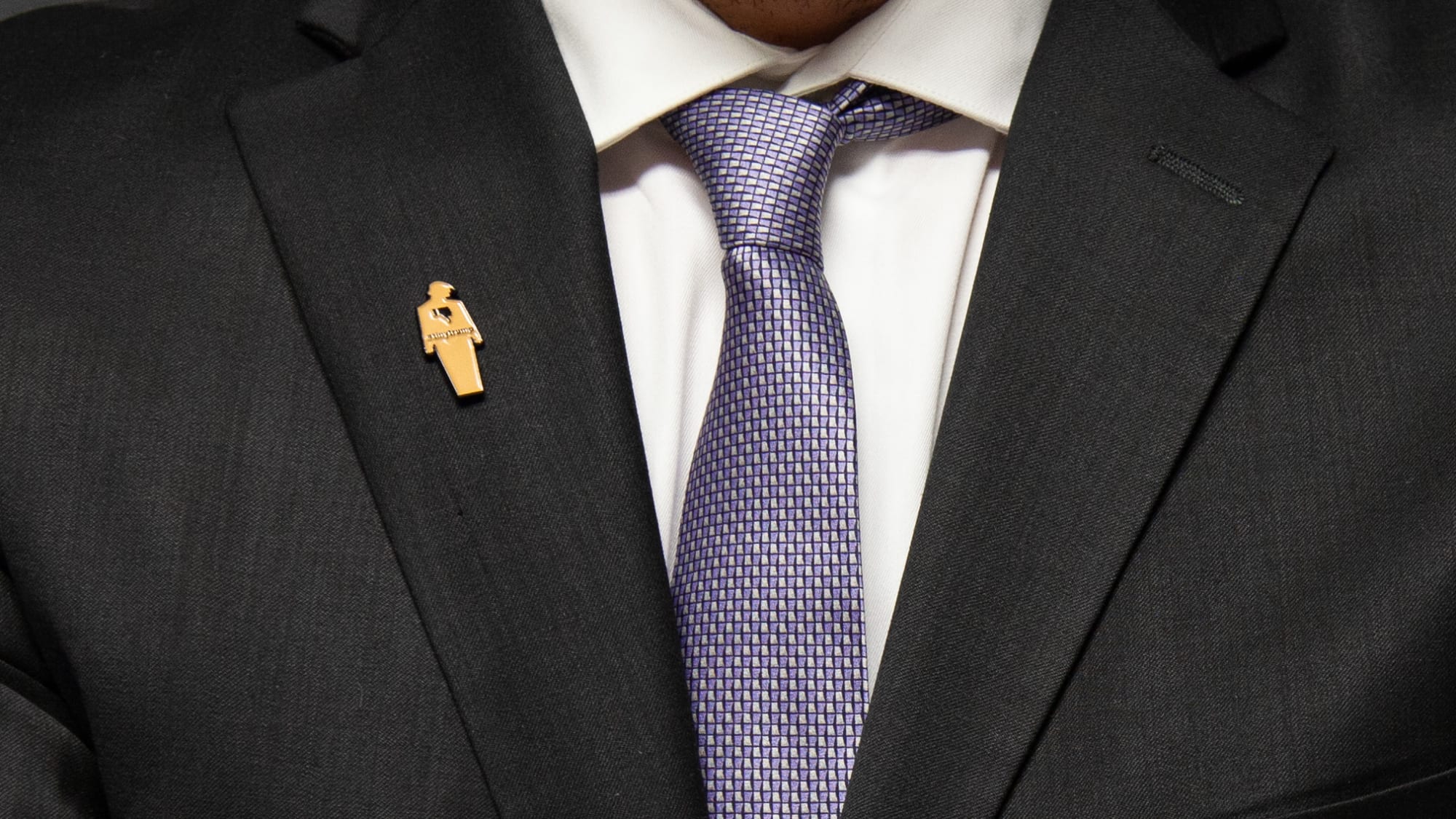 Photograph of a brown tiny trump enamel pin with a thumbs down sign on the left lapel of a man's suit blazer