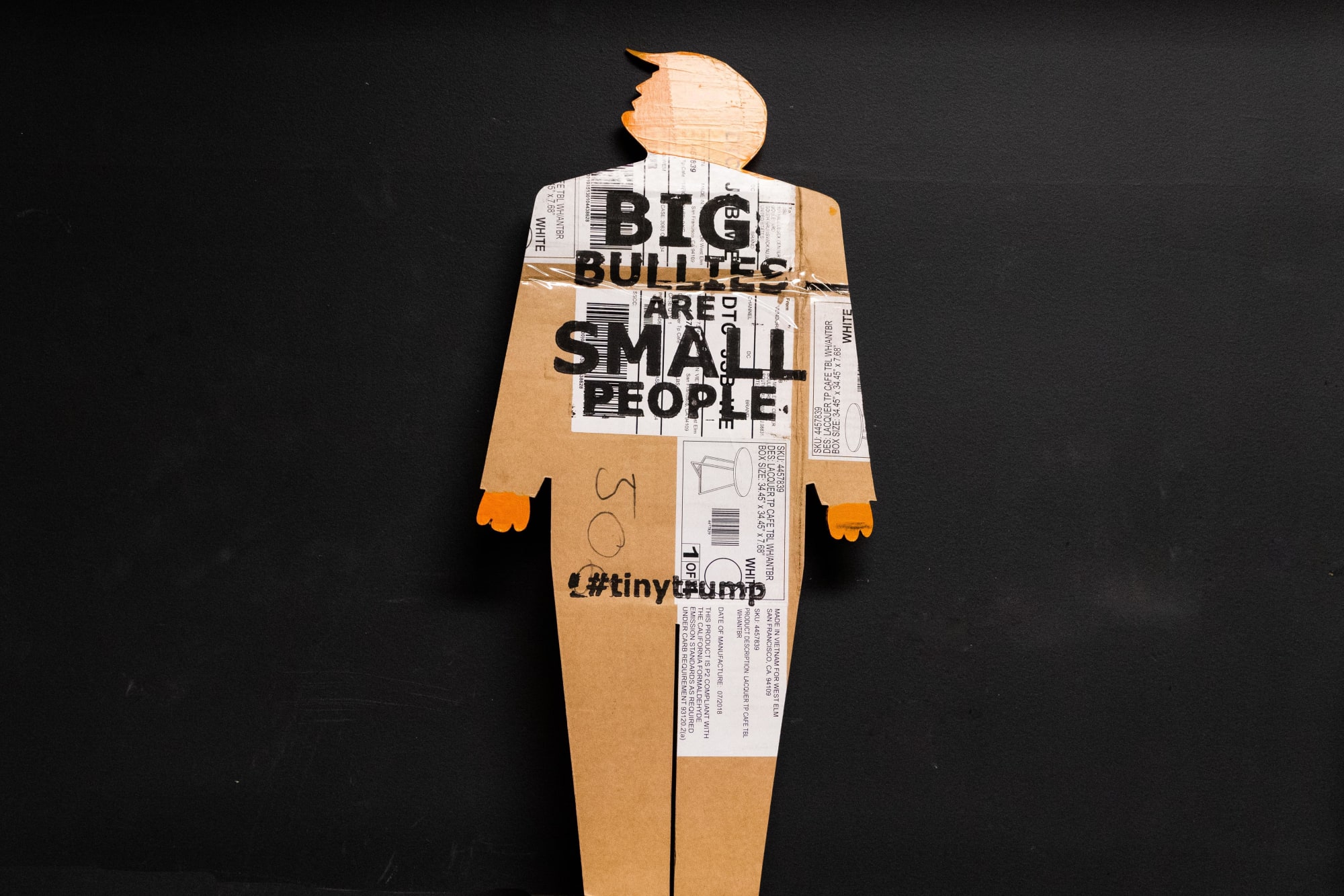 Two foot tall caricature cardboard cut out of trump with clown like hands and swooshy hair. Head and hands are painted bright orange and the slogan Big Bullies Are Small People is stamped in black ink across the chest. The cardboard is clearly used—it has shipping labels on it. Gray photo studio background.
