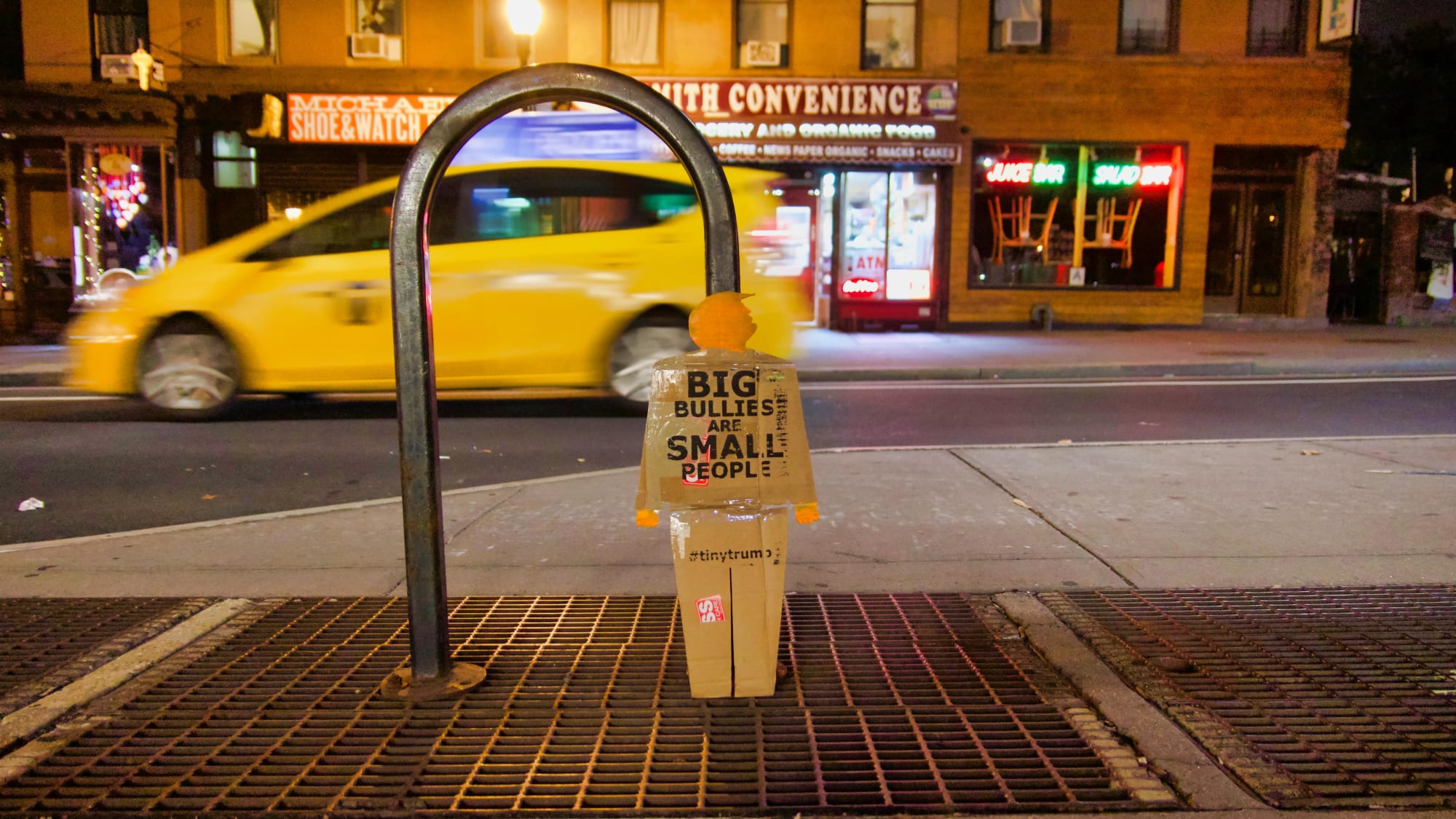 A two foot tall cardboard cutout tiny trump protest sign (in the image of a full bodied trump) leaning against a bike rack in the street in Brooklyn, New York. It is nightime and a yellow cab is blurred in the background as it whizes by.