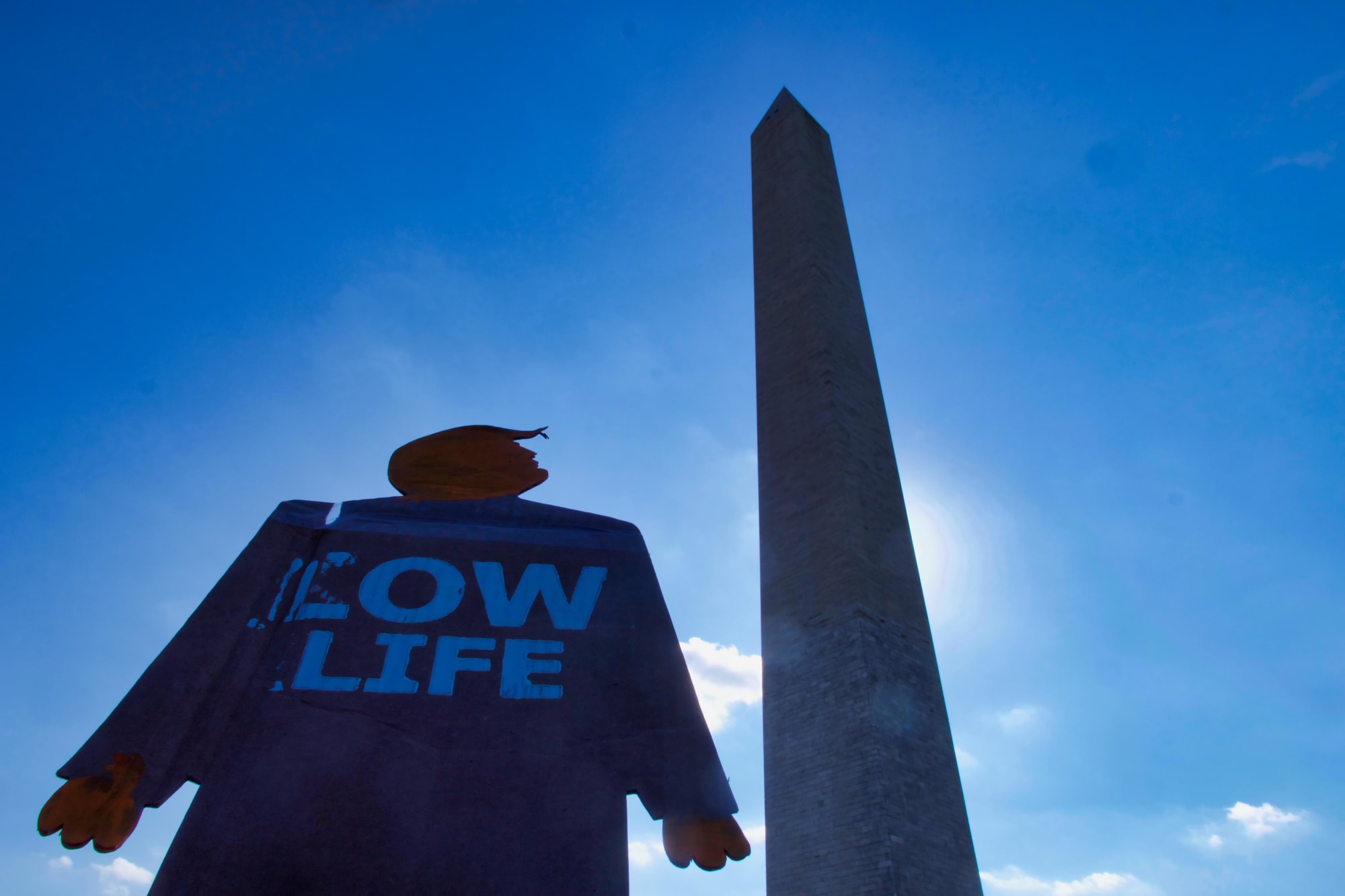 A two foot tall cardboard cutout tiny trump protest sign with the slogan Low Life looking up at the Washington Monument in Washington, D.C. Photograph angle is pointed up sharply so only the tiny trump, monument, and sky are visible