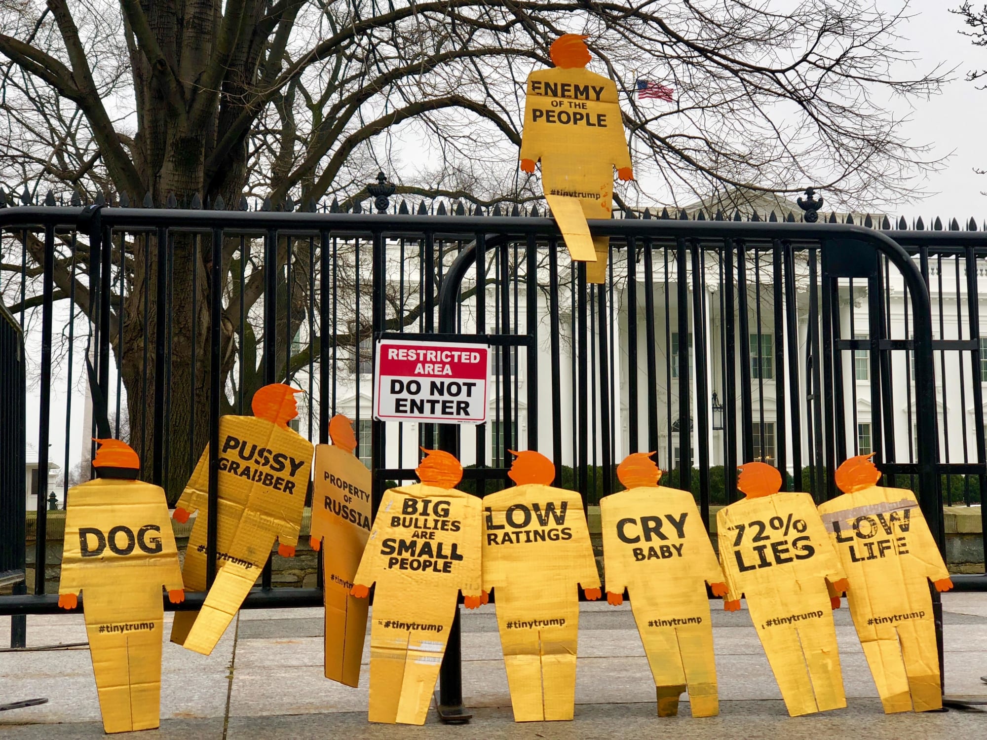 12 golden tiny trumps standing on their own accord against a gate outside of the White House