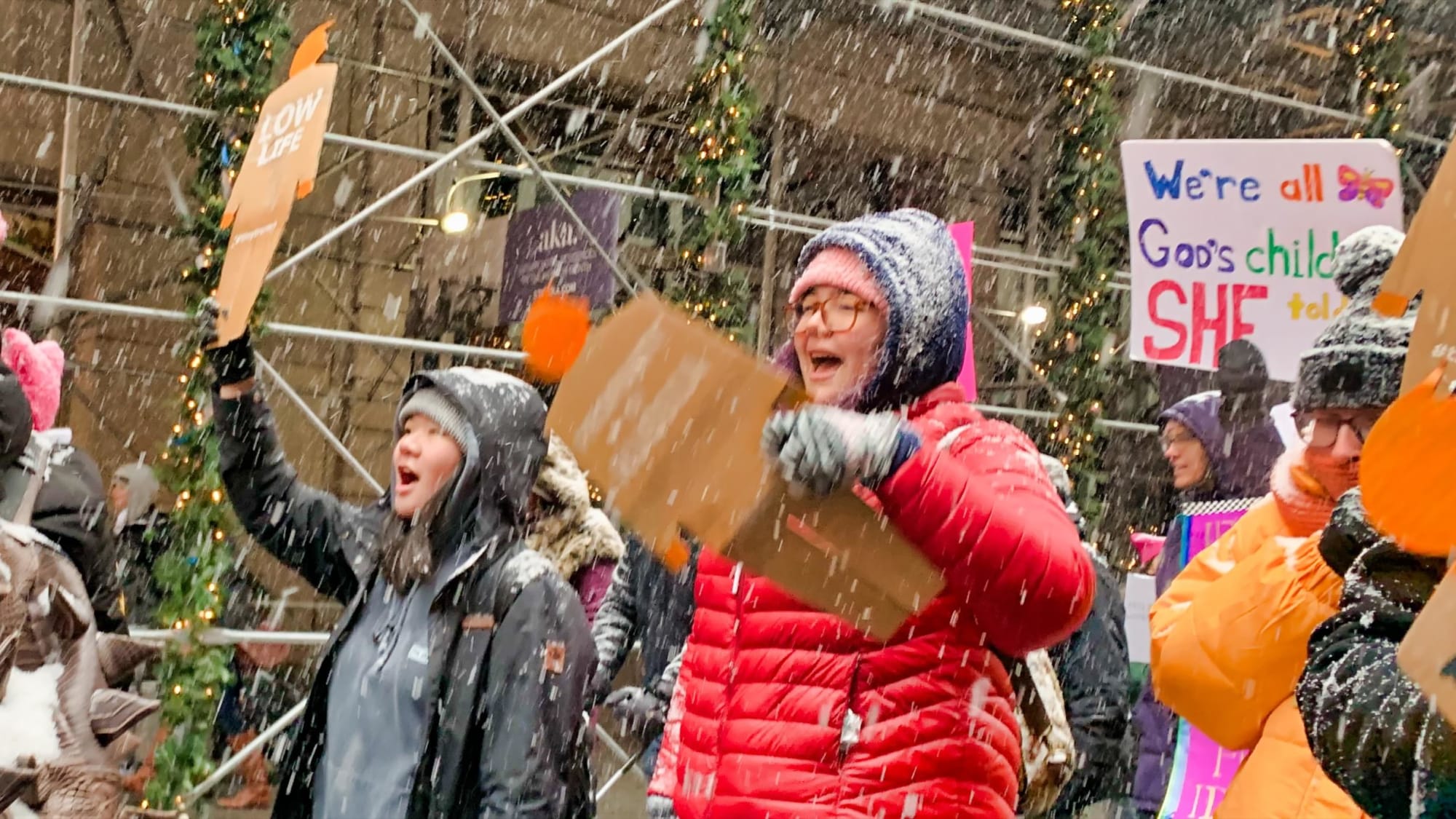 Photograph from the side of two students marching through a very windy and snowy New York City street, each holding a tiny trump