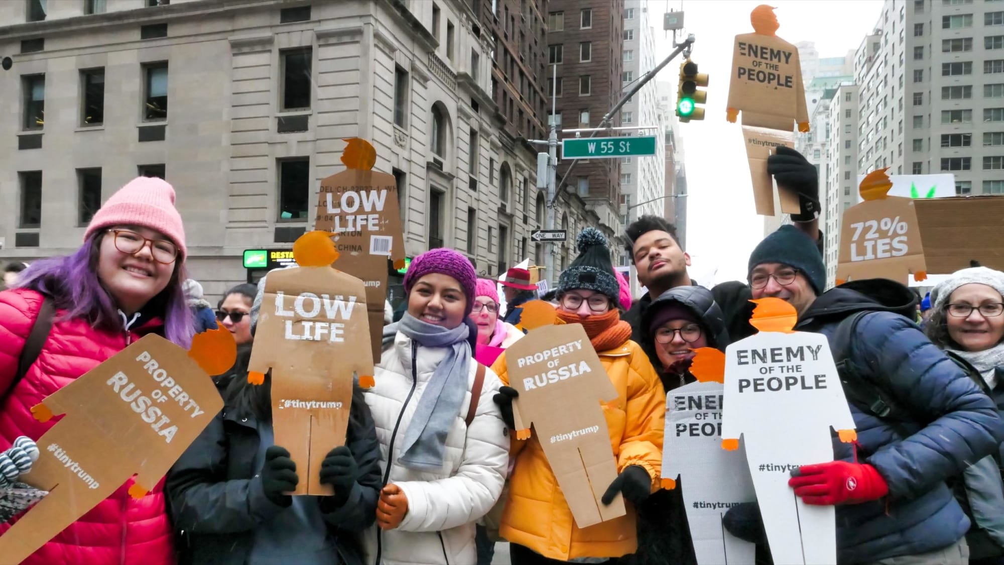 Group photograph 8 people, including the artist, holding up tiny trumps during the protest march, in a NYC intersection. In the background the street sign denoting 55th street is visible.