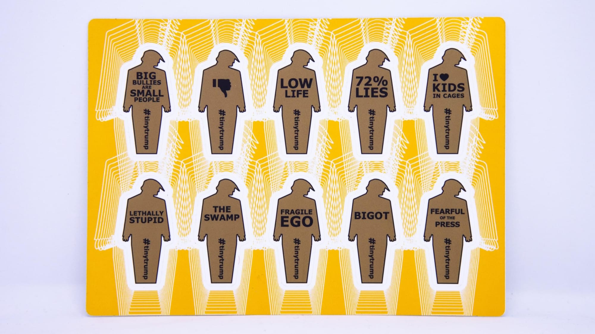 Small thumbnail image of Product photograph of a 4 inch by 6 inch sticker sheet consisting of a yellow background and 12 individual tiny trumps with the following slogans: Big Bullies Are Small People, Thumbs Down, Low Life, 72% Lies, I love kids in cages, Lethally Stupid, The Swamp, Fragile Ego, Bigot, Fearful of the Press