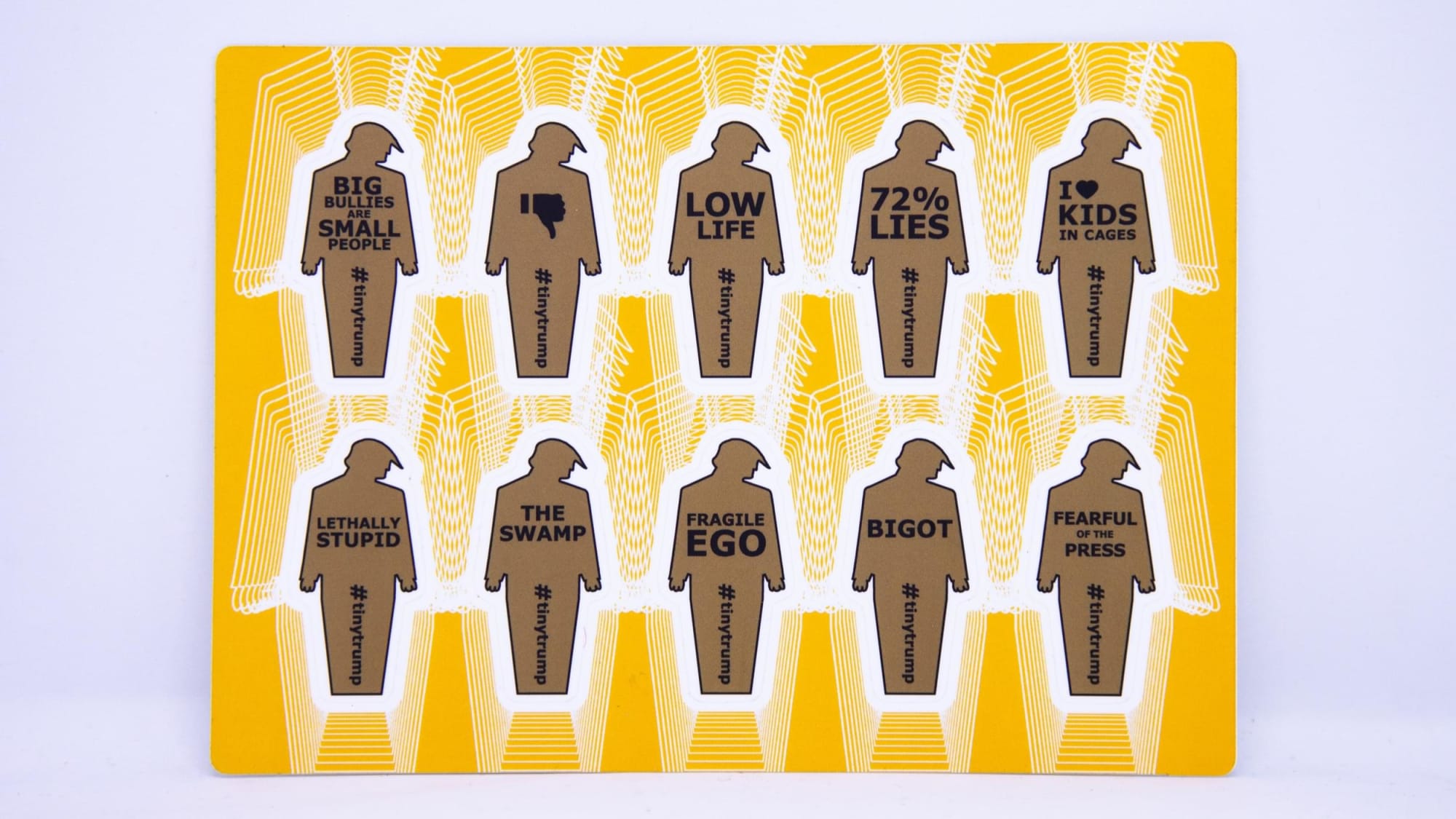 Product photograph of a 4 inch by 6 inch sticker sheet consisting of a yellow background and 12 individual tiny trumps with the following slogans: Big Bullies Are Small People, Thumbs Down, Low Life, 72% Lies, I love kids in cages, Lethally Stupid, The Swamp, Fragile Ego, Bigot, Fearful of the Press