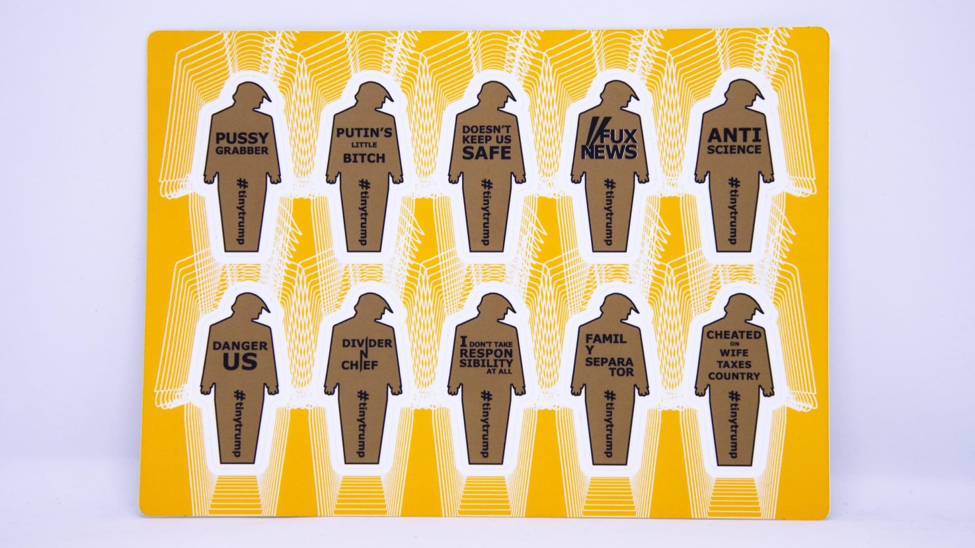 Product photograph of a 4 inch by 6 inch sticker sheet consisting of a yellow background and 12 individual tiny trumps with the following slogans: Property of Russia, American Virus, Con & Troll, Dictator, Sexist, Low Ratings, Sad, Lies Lies Lies, Unfit, Black hole graphic