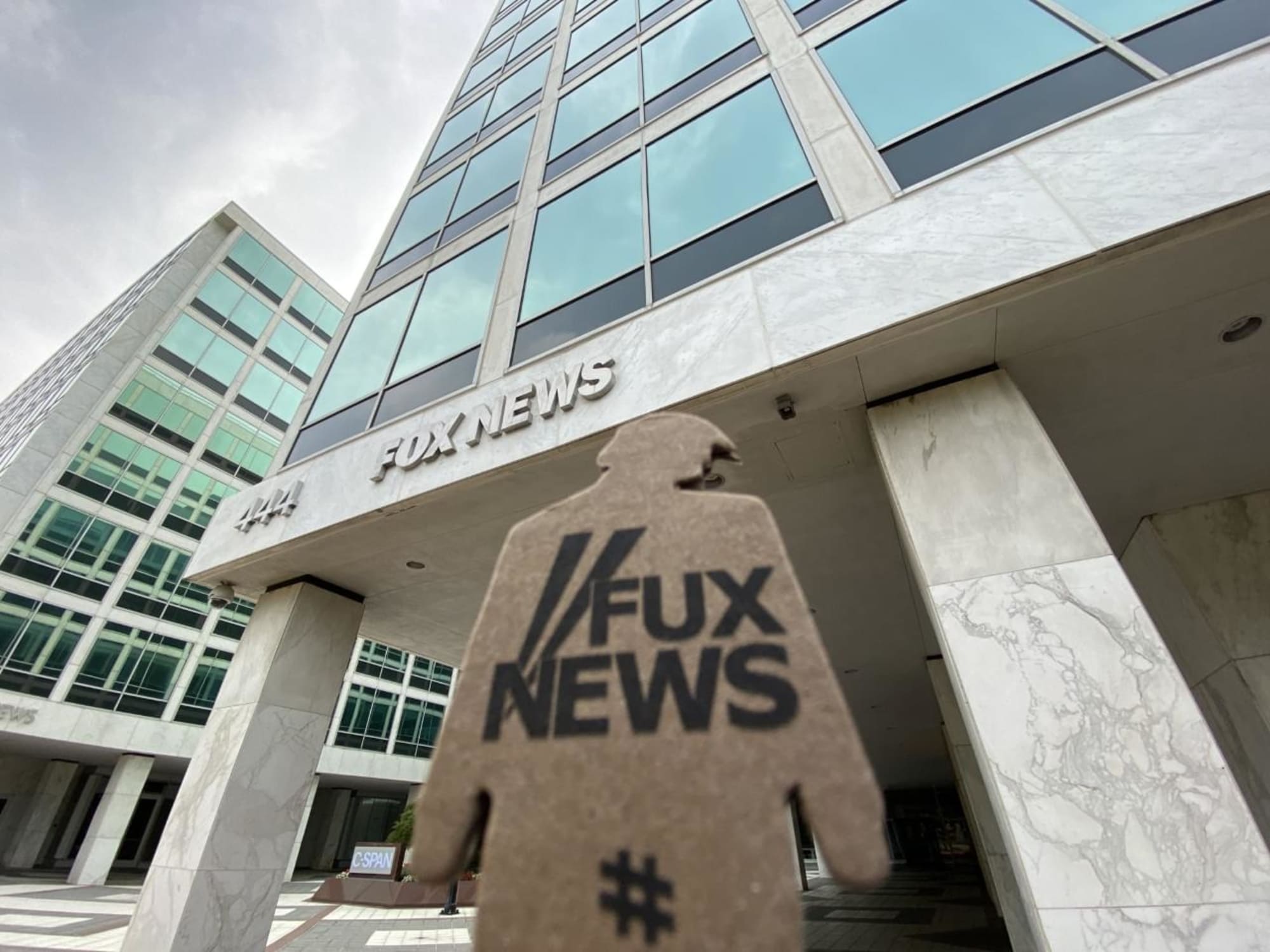 a photograph of a tiny trump with the slogan 'Fux News' held in front of Fox News headquarters in NYC