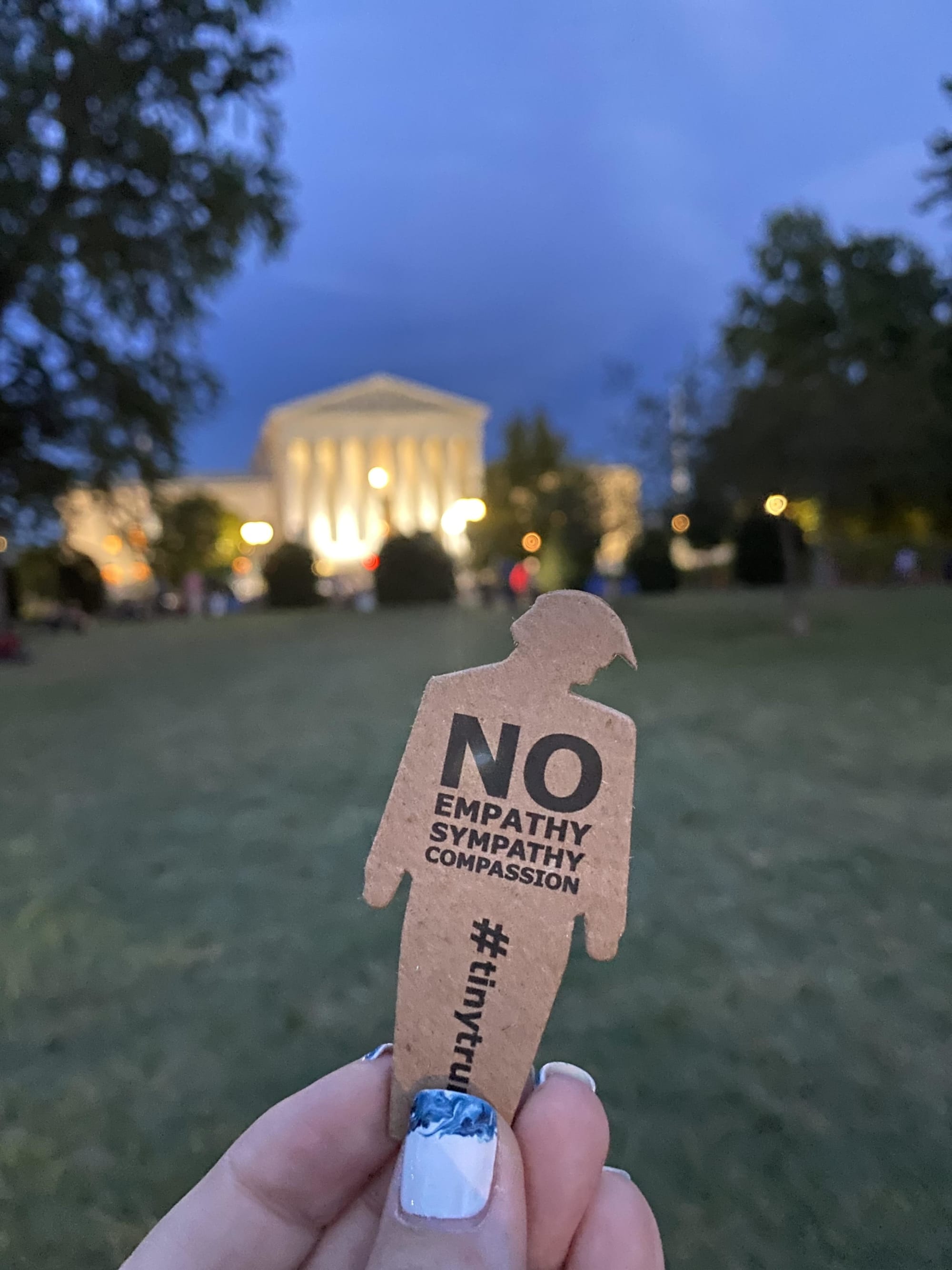 a photograph of someone holding a tiny trump with the slogan 'No Empathy, No Sympathy, No Compassion' in front of the White House