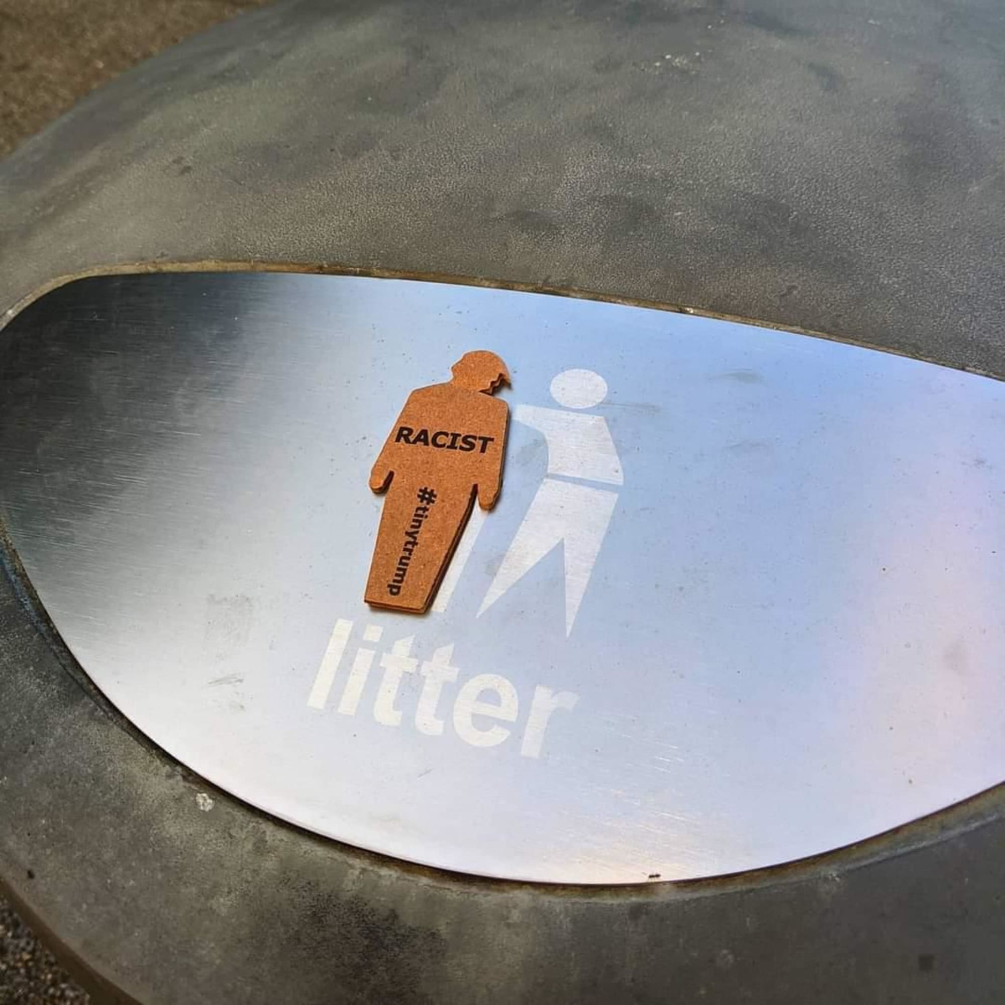 a photograph of a tiny trump with the slogan 'Racist' stuck to a garbage can that's labeled 'Litter'