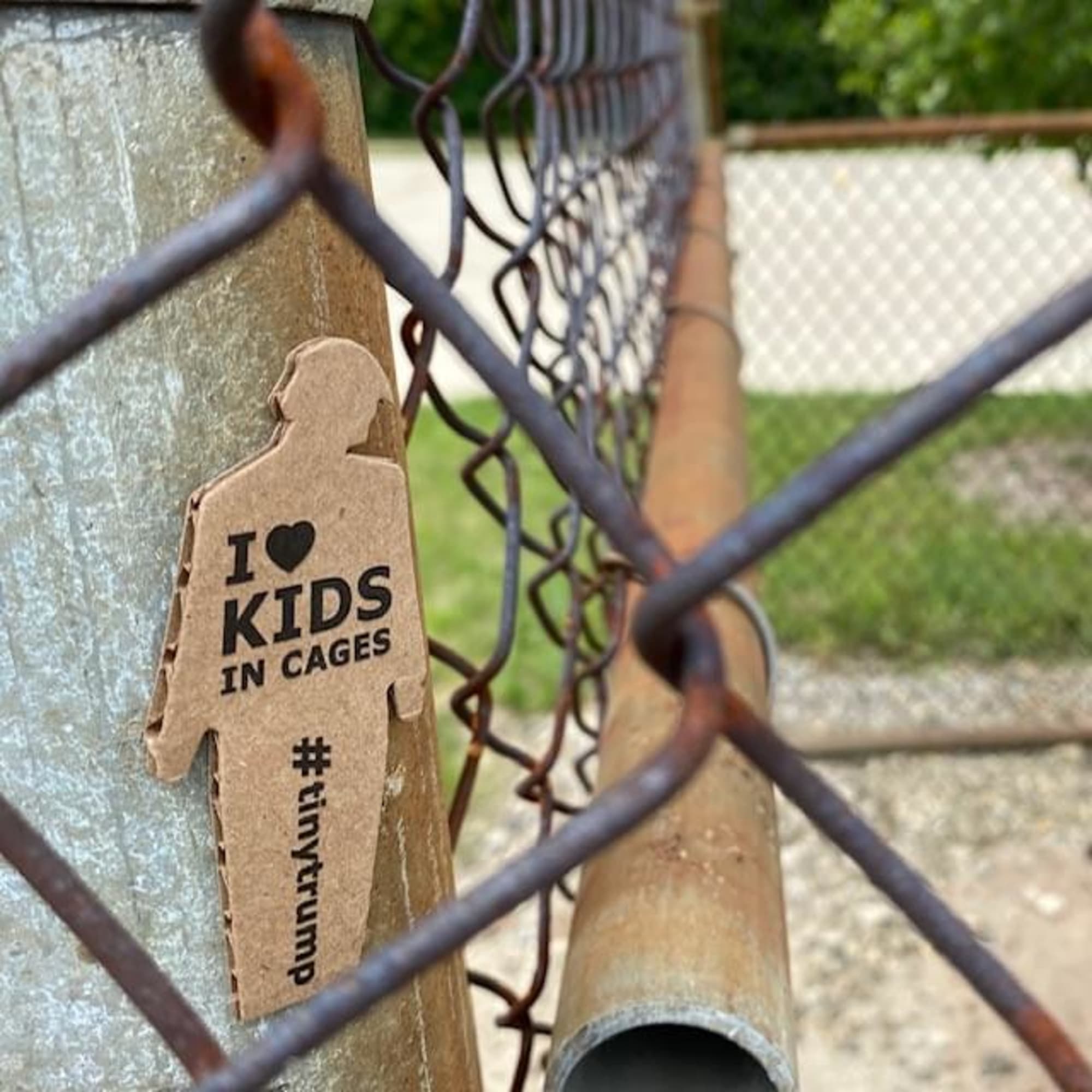 a photograph of a tiny trump with the slogan 'I love kids in cages' shown stuck to the post of a chain link fence