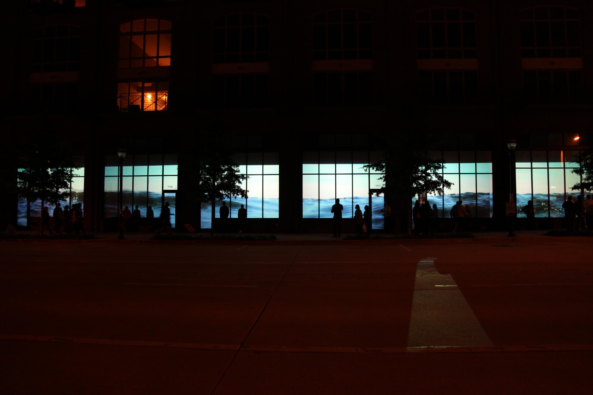 Nighttime photograph of a large commercial center with six of its street-level storefront windows showing a contiguous video projection of water ebbing and flowing.