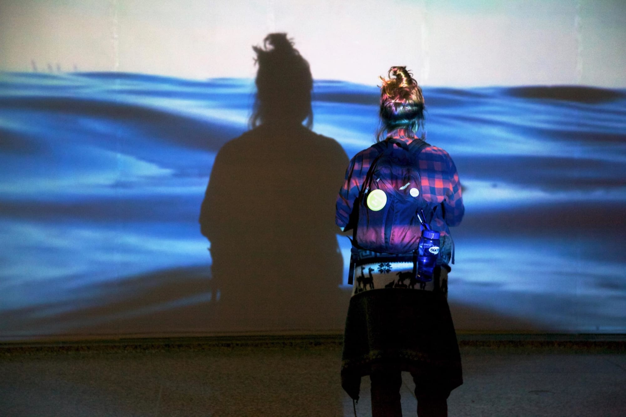 Photograph of the back of a seemingly teenage girl wearing a backpack staring at the life-size projection of water in front of her. We see her shadow in the projection of the water.