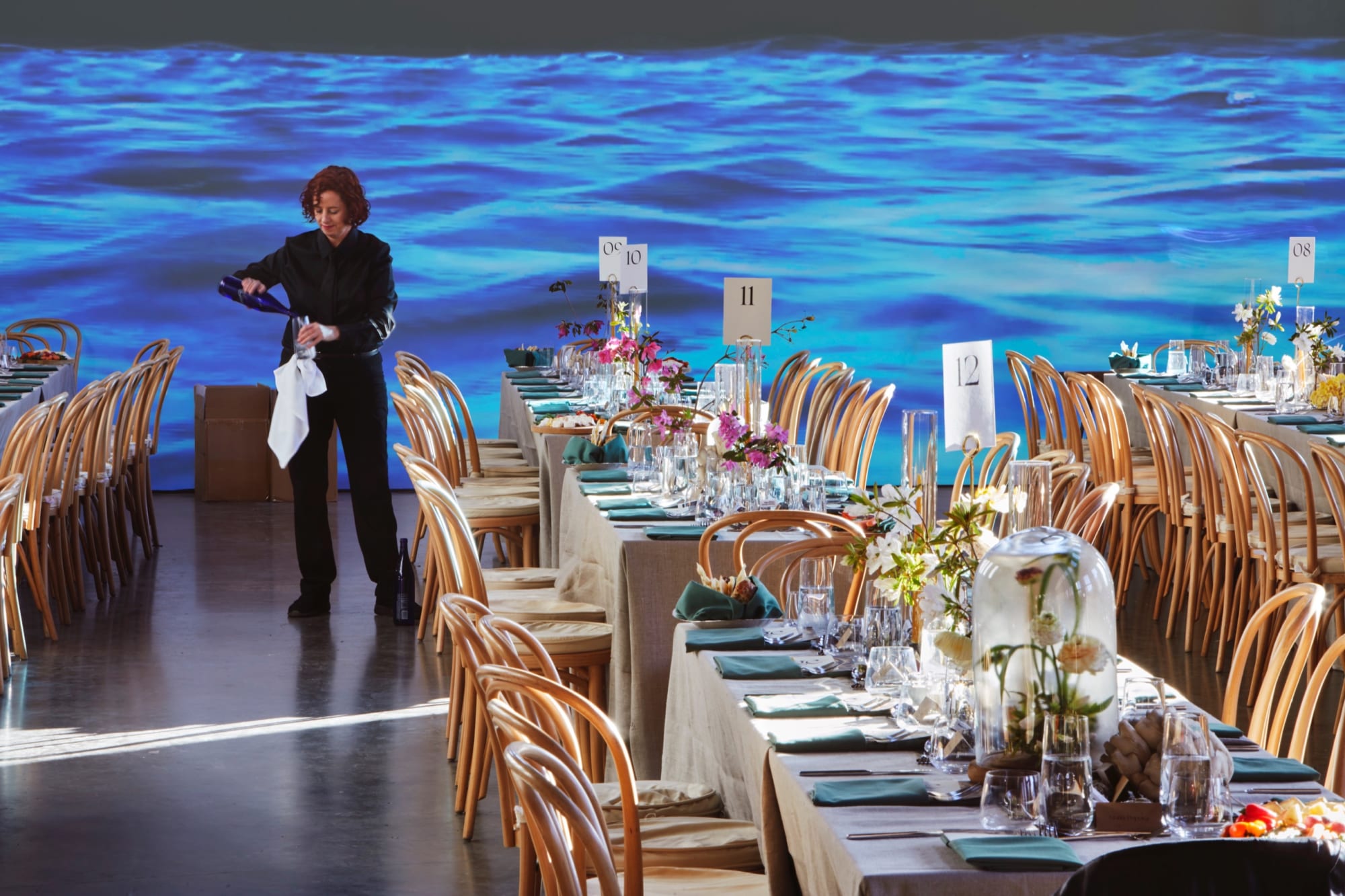 Foreground: white table clothed tables with numbers suggesting a gala event, though no one is seated at them yet. Middleground: a server pours water into a glass. Background: a video of a body of water is projected on a white wall.