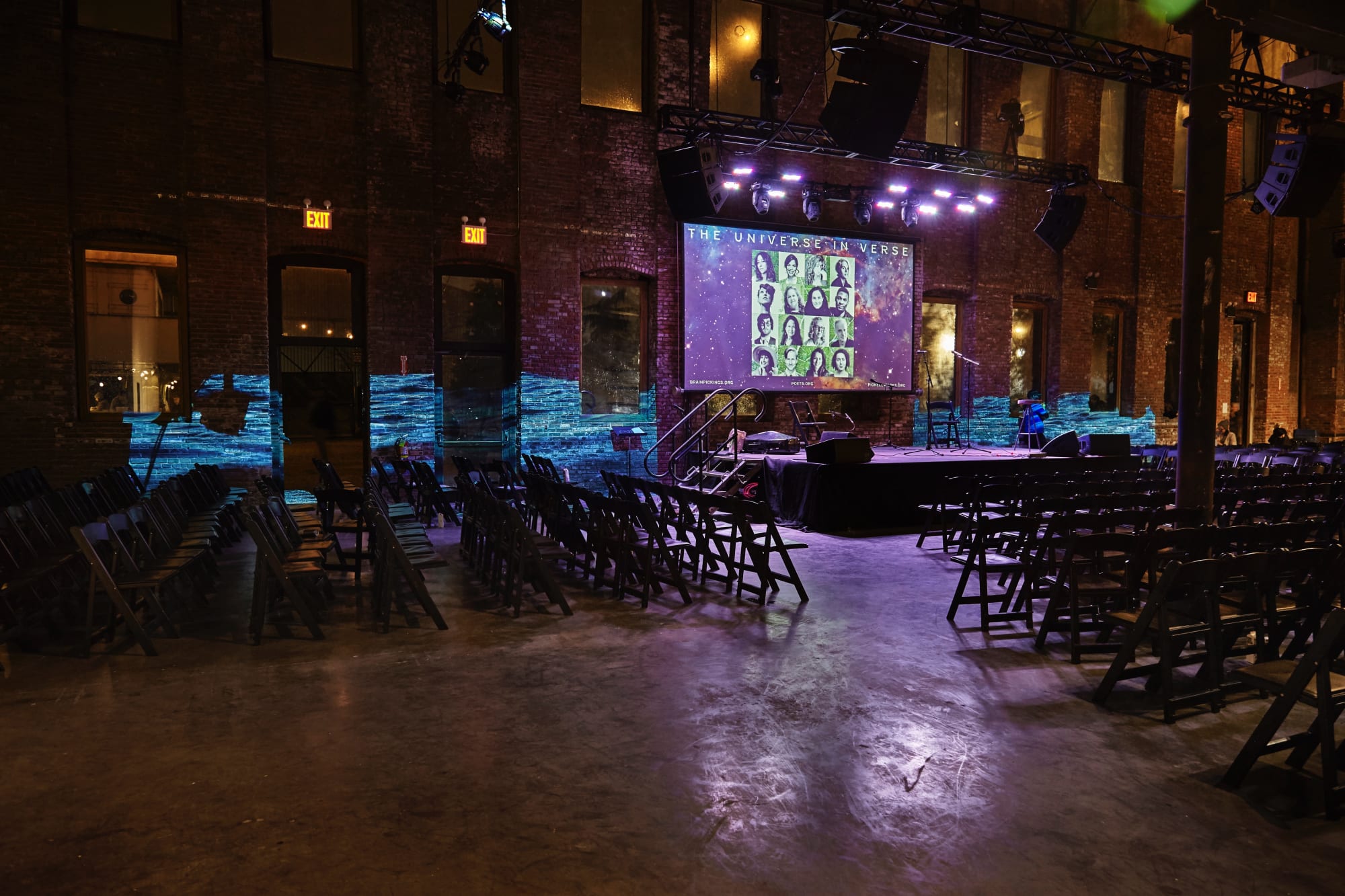 Photograph of a large empty room with over a hundred empty folding chairs. A small stage is in the center and above it a screen indicates the name of the event: 'The Universe In Verse.' A video of a body of water is projected on the brick wall in the background.