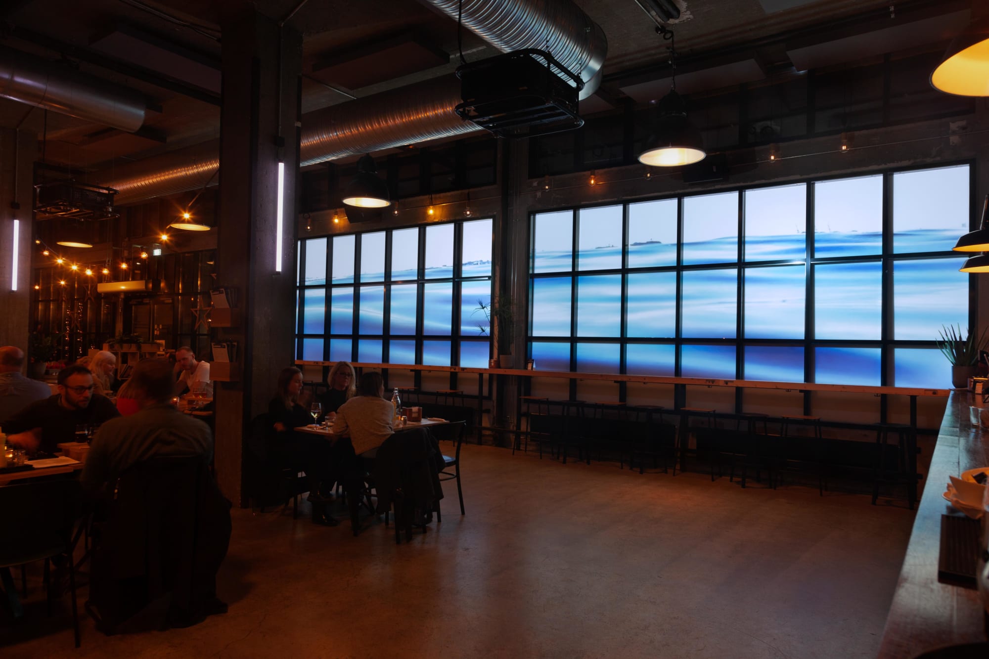 Interior photograph showing people at a bar doing normal bar stuff with Water Will Be Here projected in the windows behind them