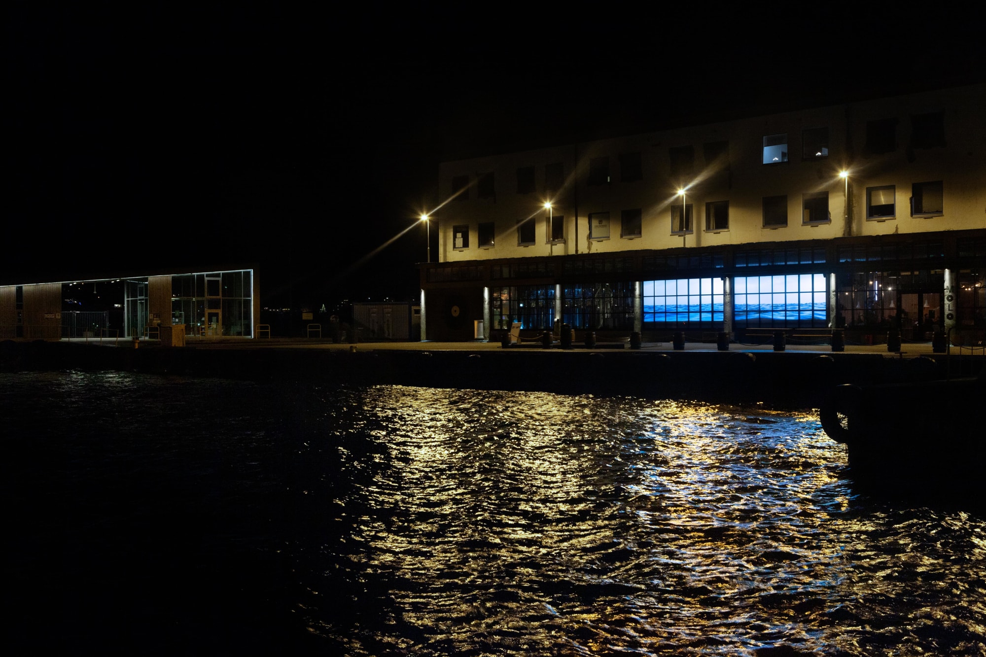 Wide shot showing Water Will Be Here (same installation as above) projected into windows of a bar about 150 feet away on the right. On the left is a body of water that is very close to the bar.