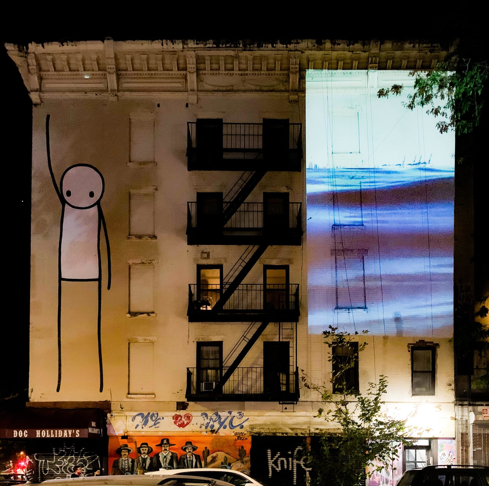 Photograph of a white building in New York City which has a 35 foot tall graffiti drawing of a stick figure reaching towards the sky on the left and Water Will Be Here projected, equally as tall, on the right