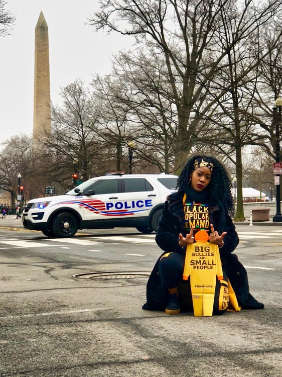 Photograph of a Black female posing with a tiny trump with the slogan Big Bullies Are Small People; she is giving the tiny trump the middle finger with both hands. In the background a police car passes by and further in the background is the Washington Monument.