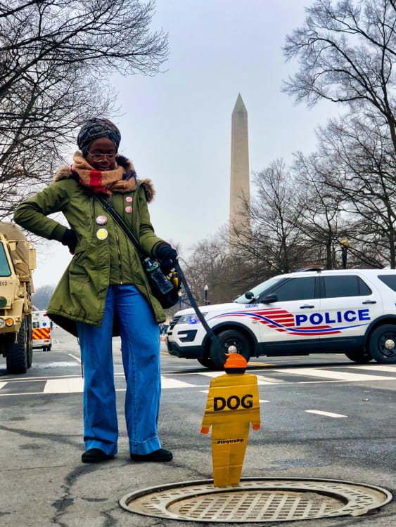 Photograph of a Black female 'walking' a tiny trump that says Dog with a dog collar. The Washington Monument is in the background.
